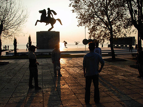Μια ομάδα Αλβανών παίζει με την μπάλα σε κύκλο και πίσω στο άγαλμα οι πιτσιρικάδες κάνουν skateboard